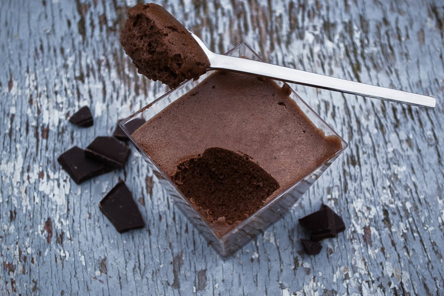 Mousse au chocolat servie dans une verrine carrée avec une cuillère et des carrés de chocolat noir sur un plan en bois gris