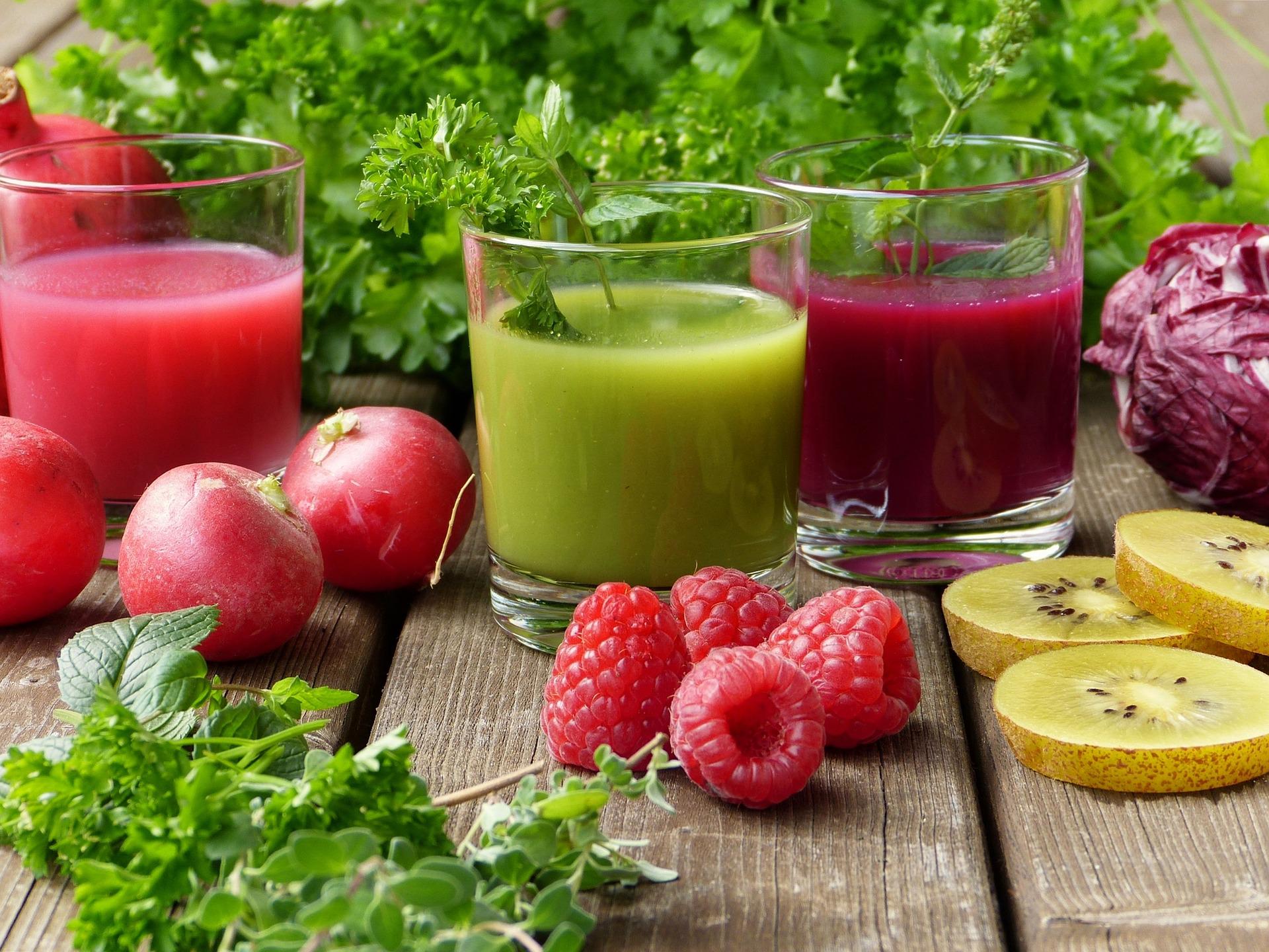 Trois verres de smoothie rose, vert et violet alignés sur une table en bois avec radis, framboises, kiwi et herbes fraîches