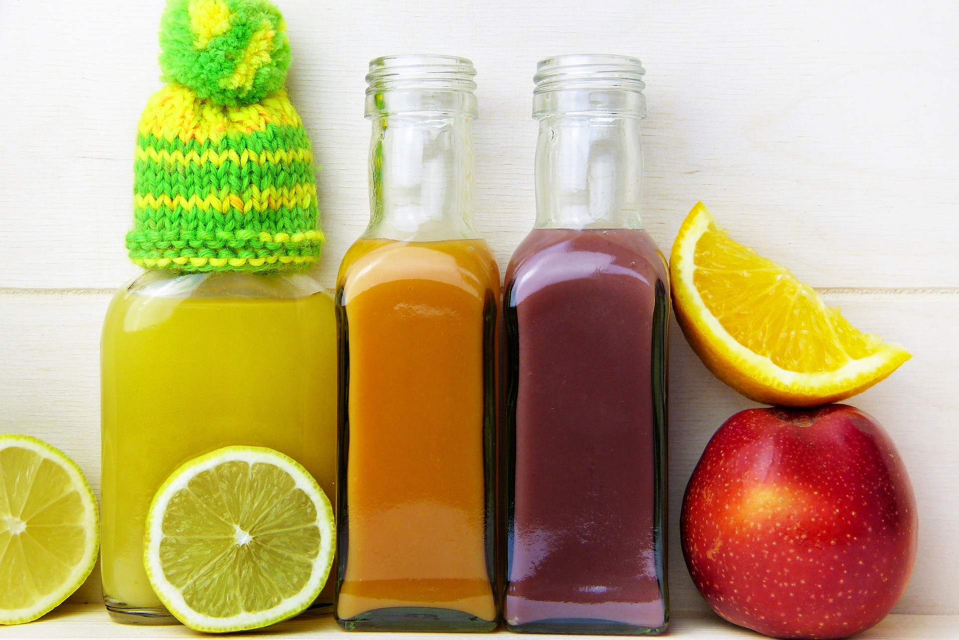 Trois bouteilles de jus jaune, orange et violet alignées entre des agrumes coupés et une pomme rouge