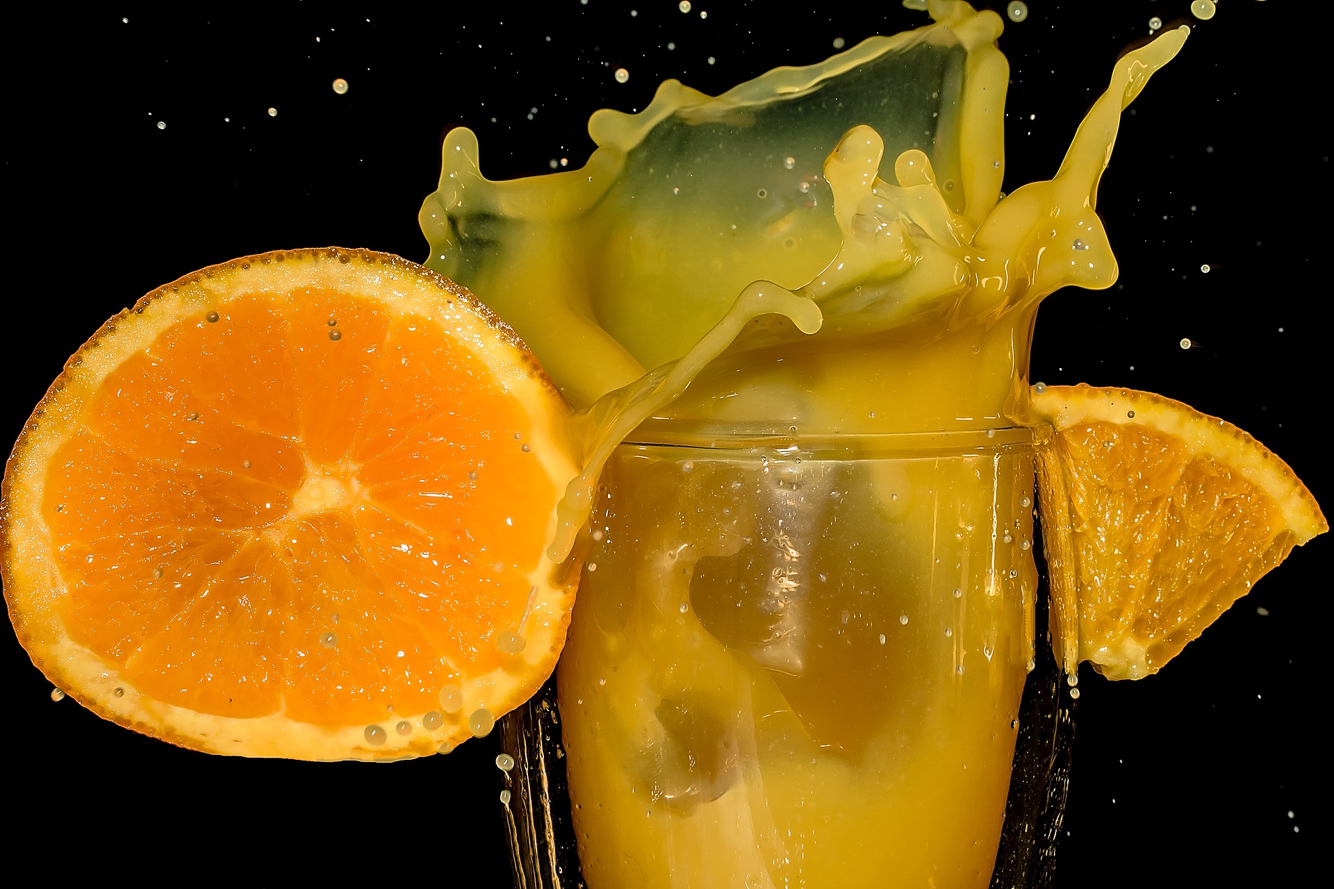 Verre de jus d'orange éclaboussant sur fond noir, avec une tranche et un quartier d'orange de chaque côté