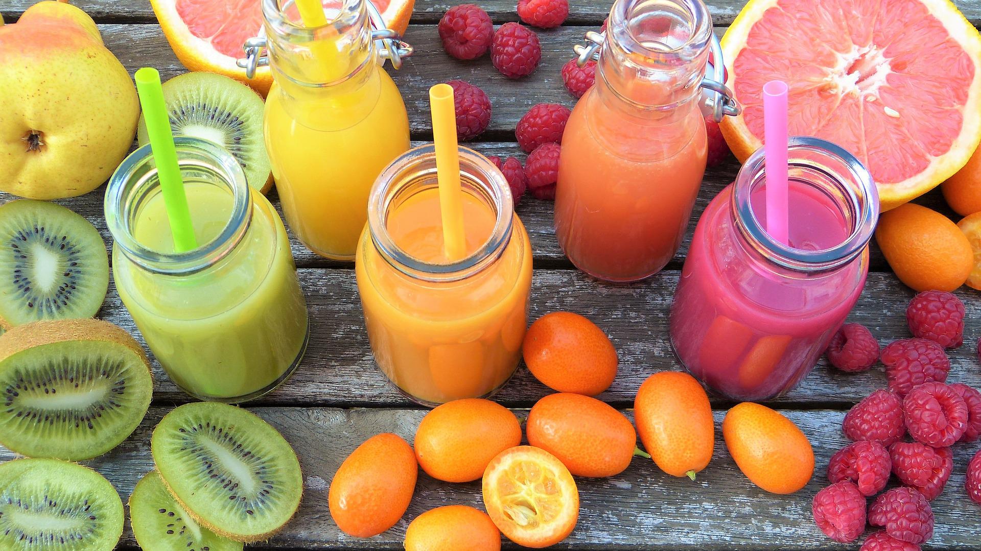 Bouteilles de smoothies épais vert, orange et rose avec pailles, posées sur des planches en bois entourées de kiwis et de framboises