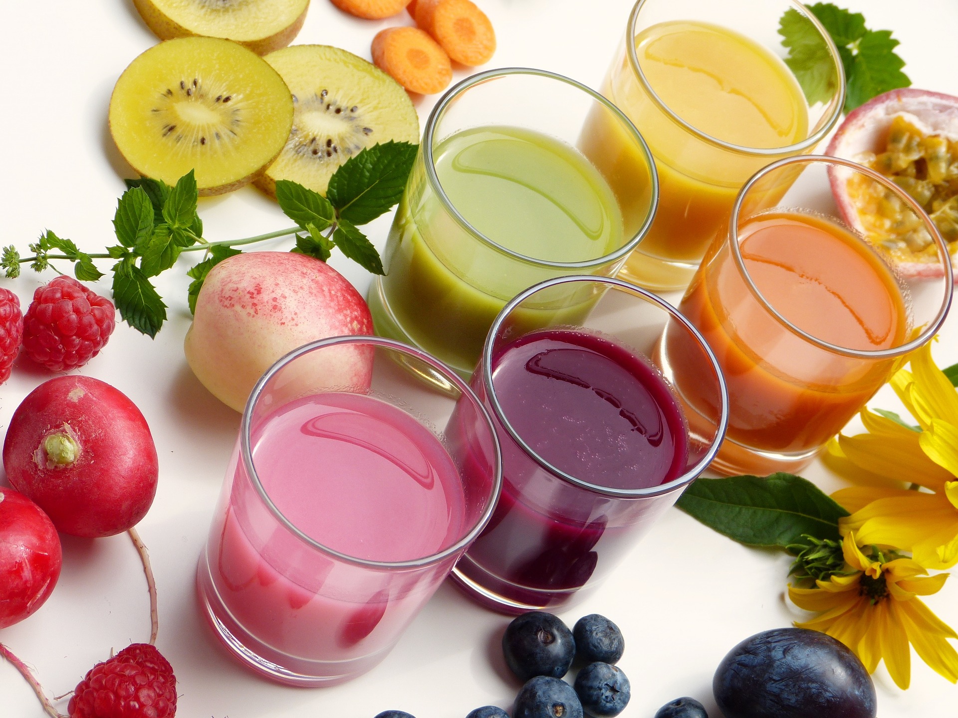 Cinq verres de jus verts, jaunes, orange et rose alignés sur fond blanc avec kiwis, carottes, framboises et fruit de la passion