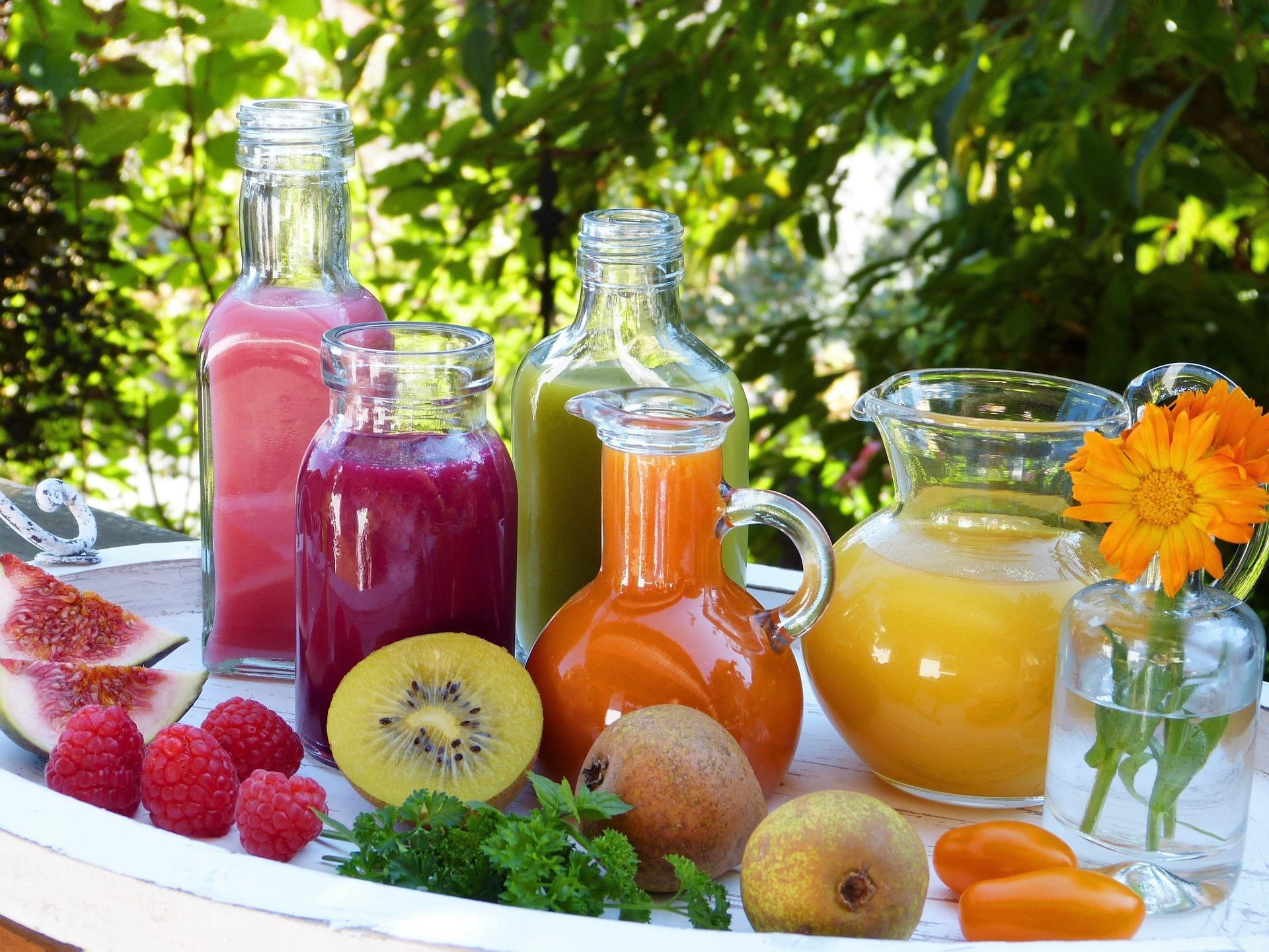 Bouteilles et carafe de jus de fruits et de légumes colorés posées sur une table de jardin blanche avec figues, poires et framboises
