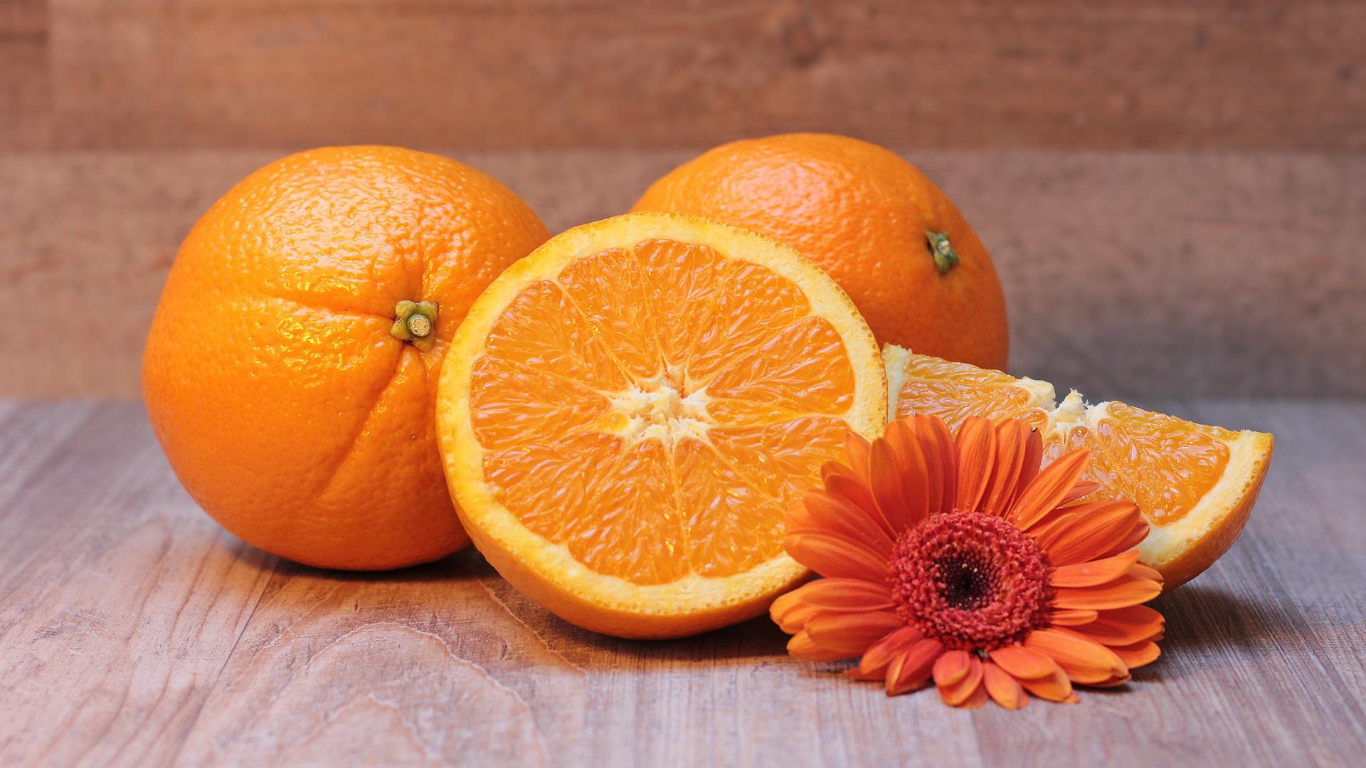 Deux oranges entières et une orange coupée en deux posées sur une table en bois clair à côté d'une fleur orange